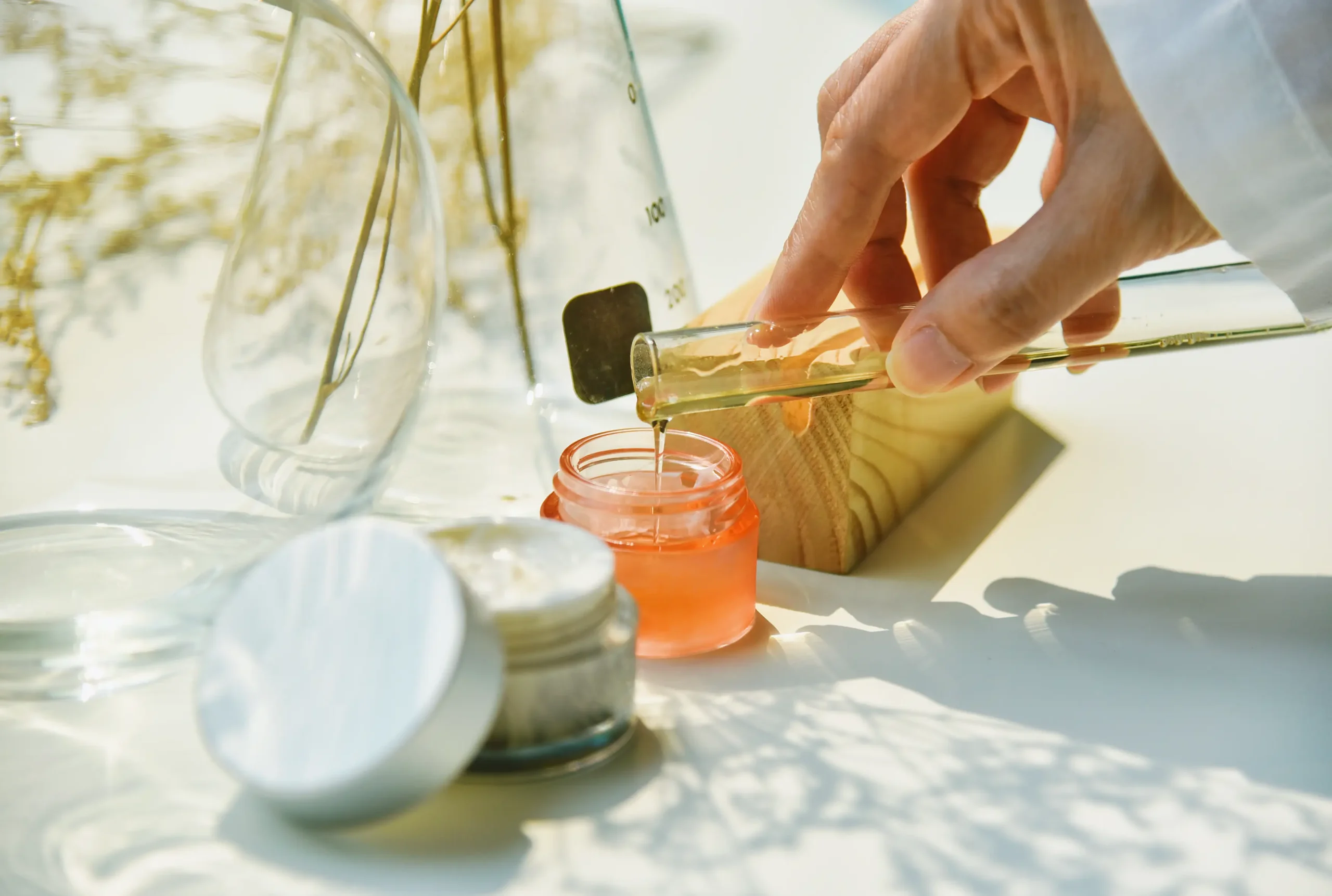 Hand pouring amber liquid from a test tube into an orange cosmetic jar, next to an open cream jar and glass beakers.