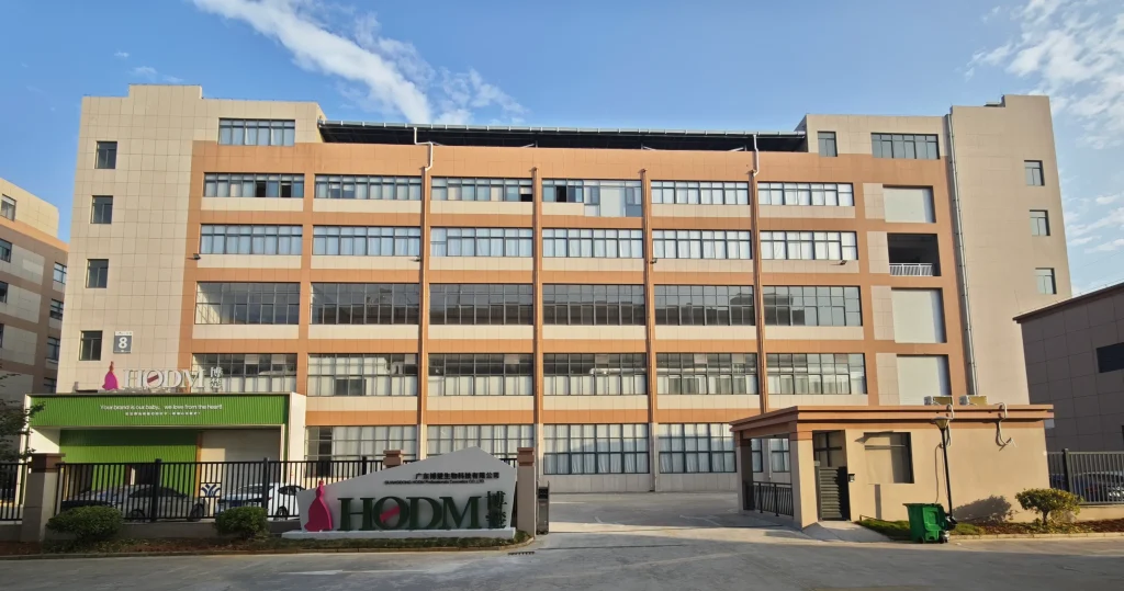 The exterior view of the HODM large multi-story factory building with a prominent company logo sign at the entrance under a clear blue sky.