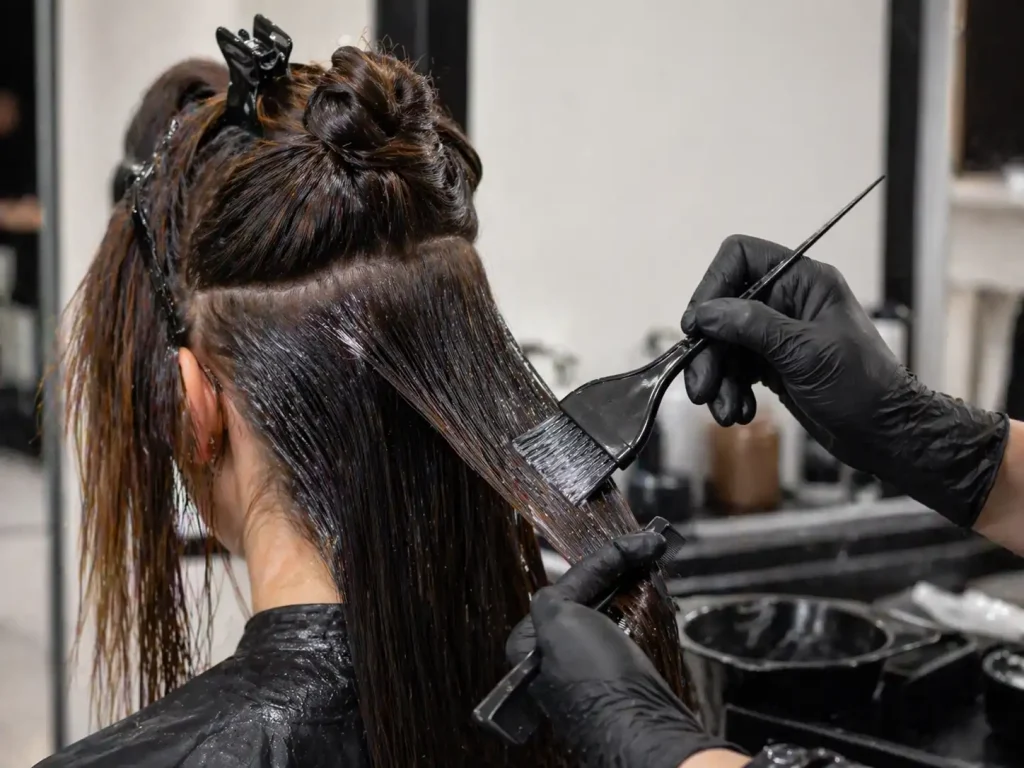 Hairdresser in black gloves applying hair dye with brushes to a client's wet, sectioned hair.
