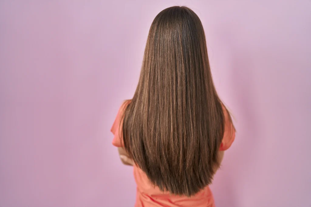 Rear view of a woman with long, smooth, shiny brown straight hair against a pink background, showing healthy texture.