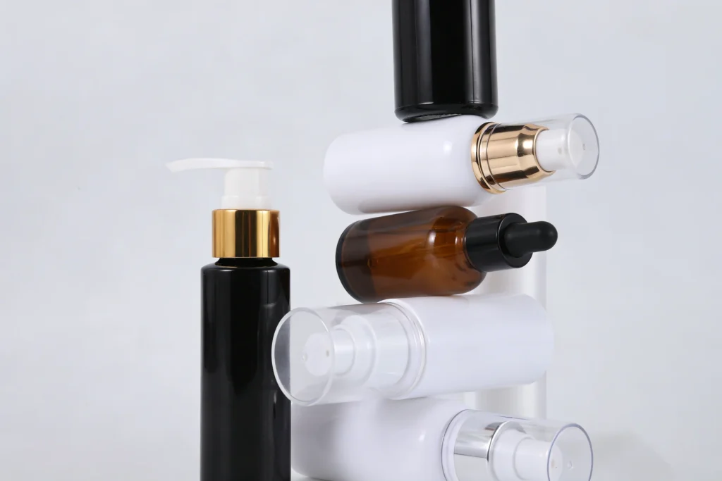 A stack of various small hair oil containers including a brown glass dropper bottle and white pump bottles, next to a tall black pump bottle with a gold collar.