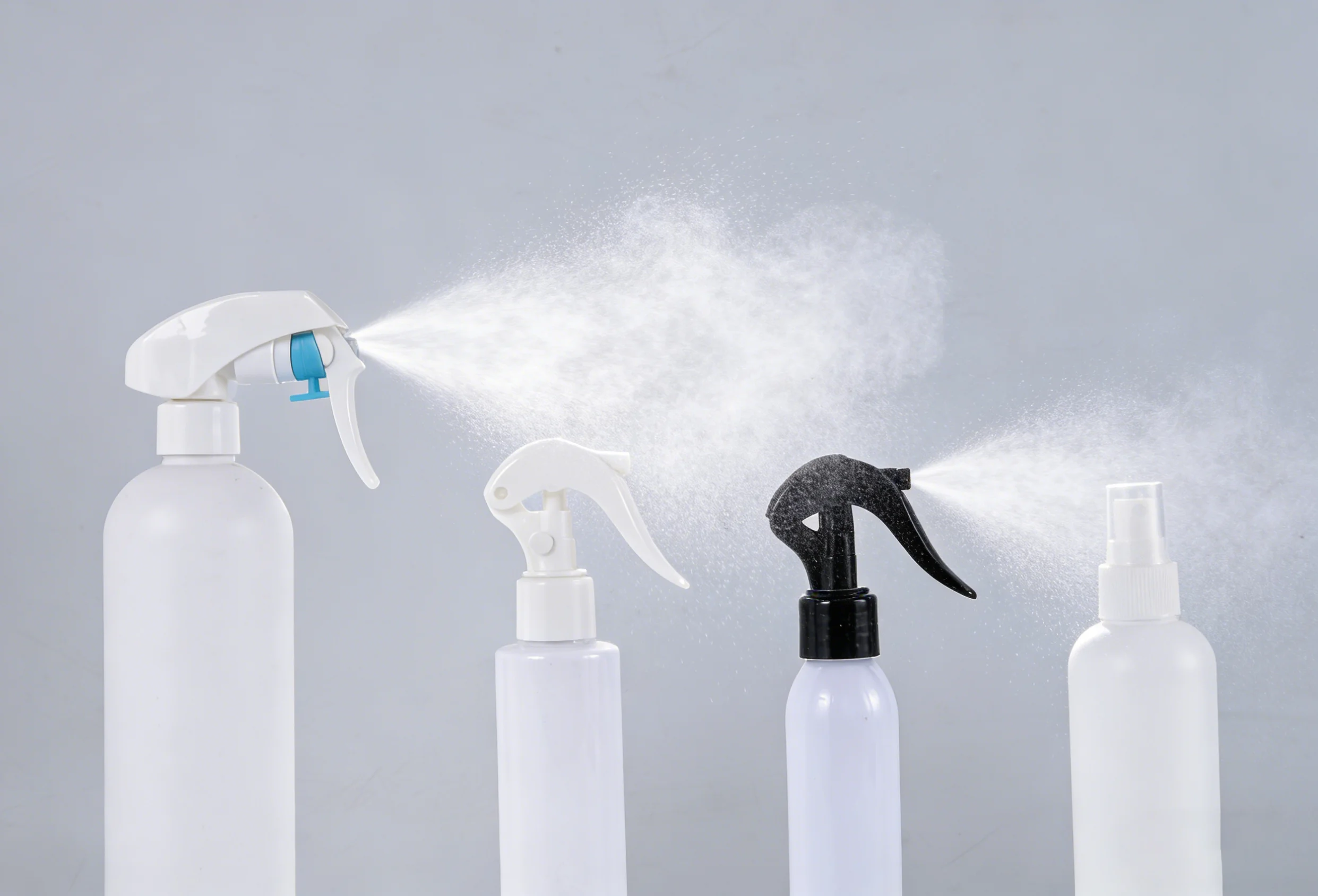 Four white spray bottles with different nozzle designs emitting fine mists against a plain grey background.