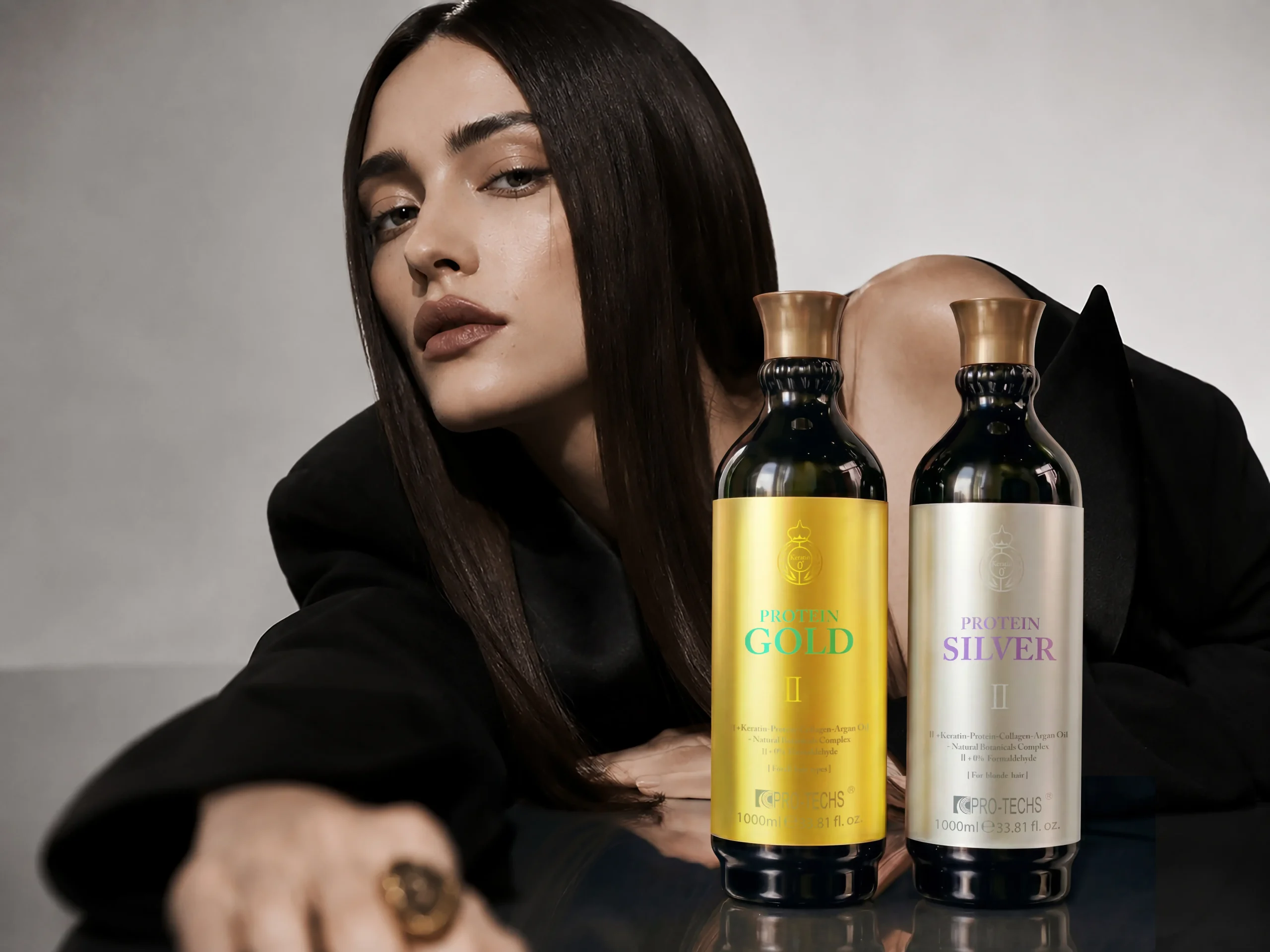 A woman with sleek dark hair posing behind large gold and silver bottles of PRO-TECHS Protein hair treatments.