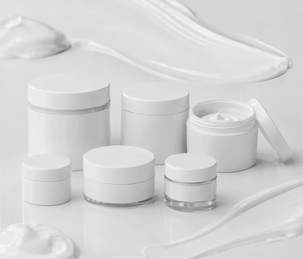 Assorted white cosmetic jars and an open jar with cream on a glossy surface, minimalistic skincare scene.