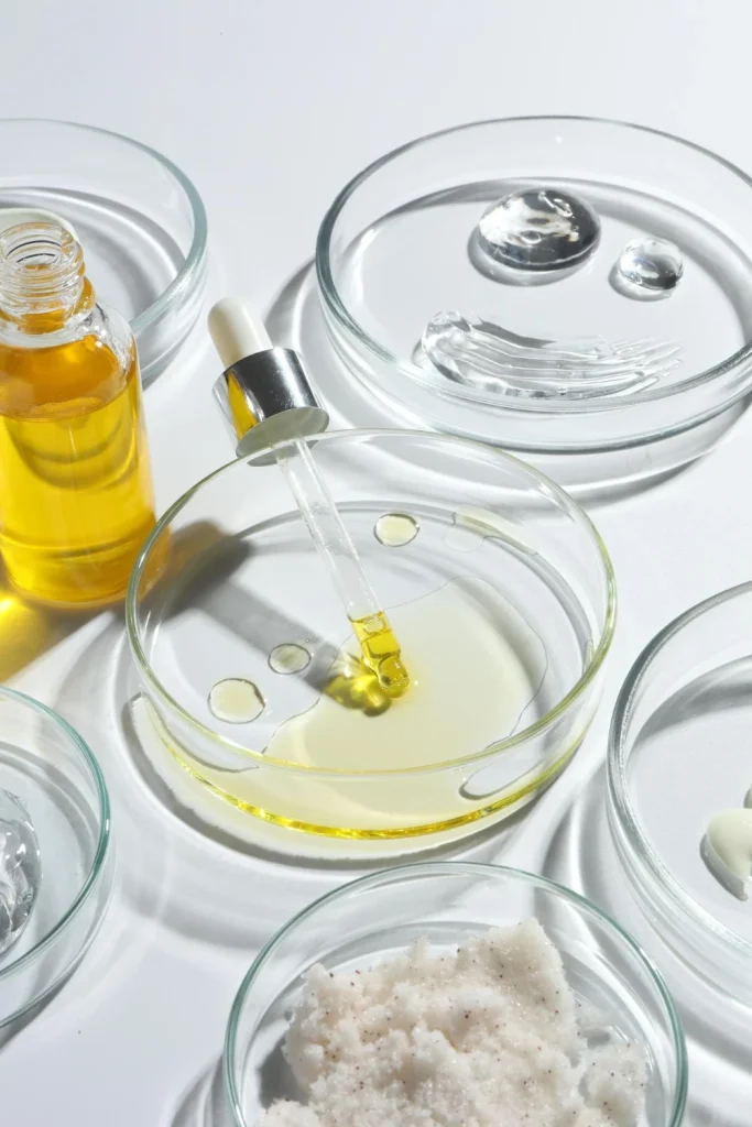 Alt="Professional hair care R&D lab setting with a dropper bottle of argan oil next to multiple petri dishes containing various cosmetic raw ingredients like clear gels, rich emulsions, and a crystalline salt scrub for custom private label formulation