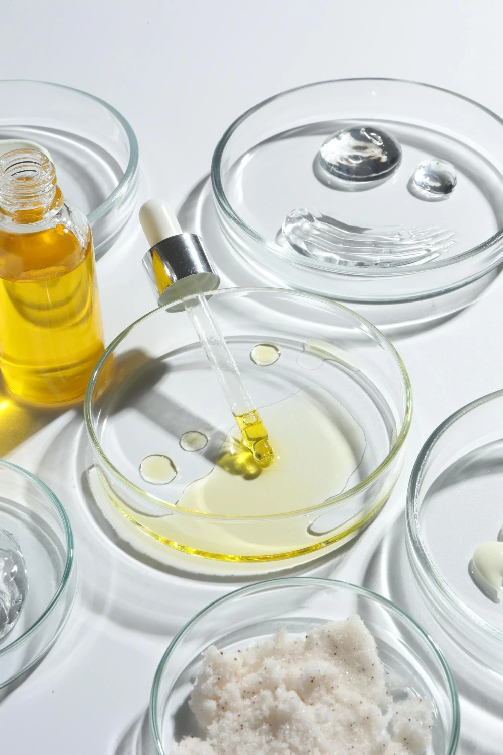 Alt="Professional hair care R&D lab setting with a dropper bottle of argan oil next to multiple petri dishes containing various cosmetic raw ingredients like clear gels, rich emulsions, and a crystalline salt scrub for custom private label formulation