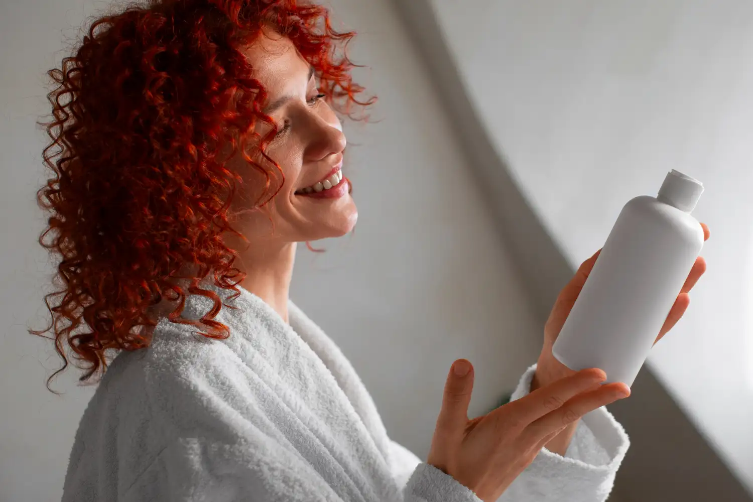 Woman with red curly hair in a white bathrobe smiles while holding a white bottle up to her face.