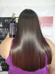 Back view of a person with long straight brown hair wearing a purple top, standing in front of hair-care banners and a pump bottle labeled keratin treatment beside their hair.