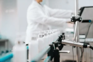 Laboratory worker in a white coat operating a bottling line with white bottles on a conveyor belt.