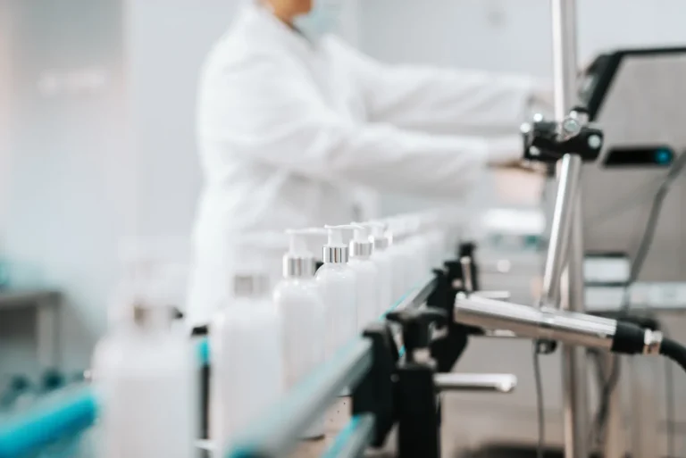 Laboratory worker in a white coat operating a bottling line with white bottles on a conveyor belt.