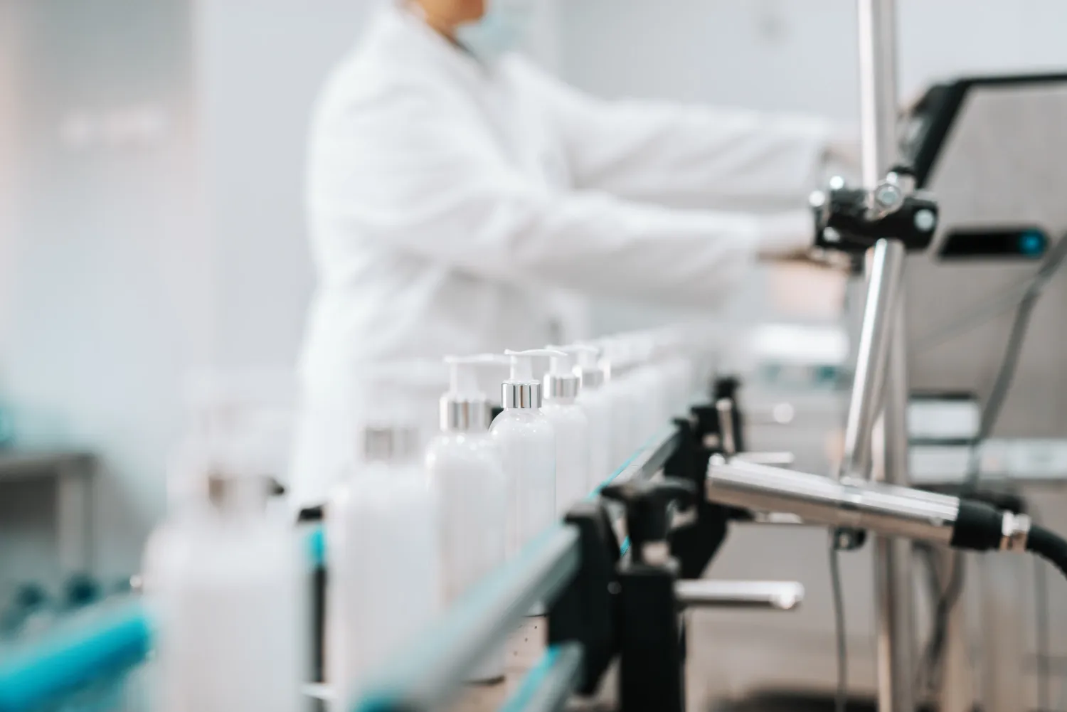 Laboratory worker in a white coat operating a bottling line with white bottles on a conveyor belt.
