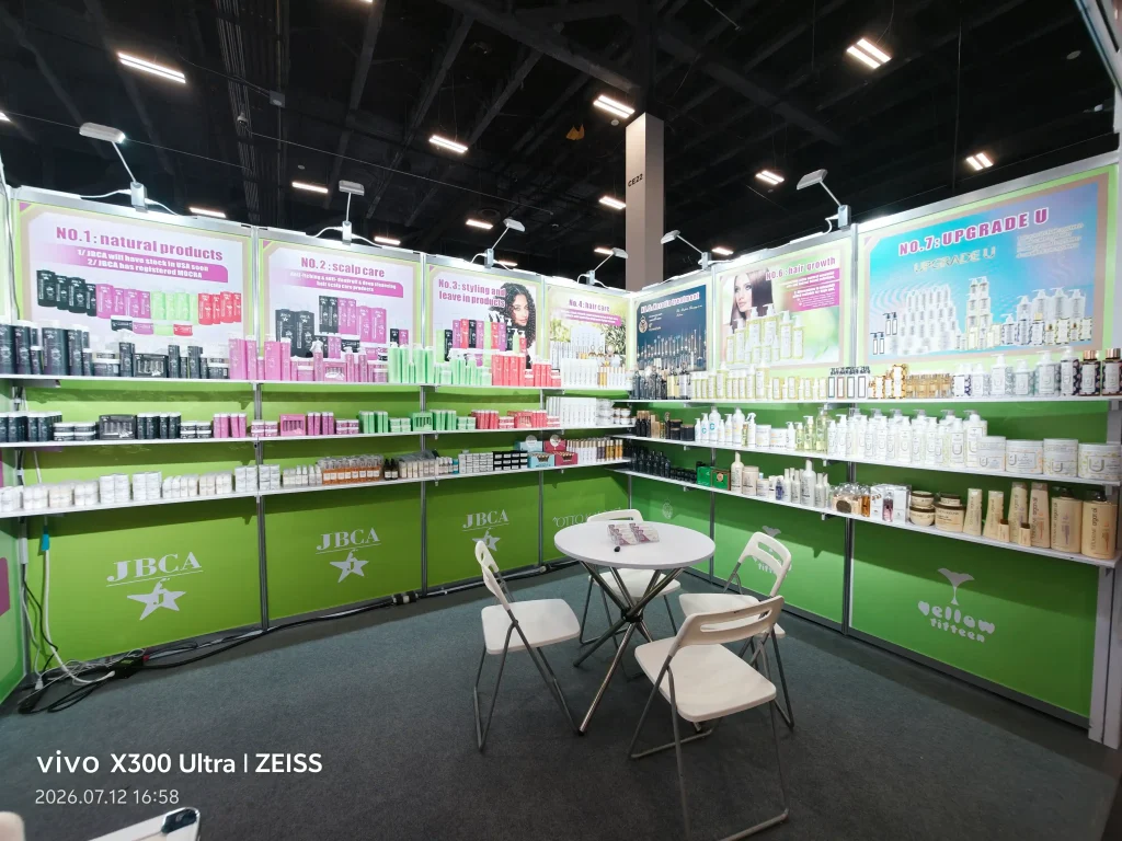 Exhibition booth with green shelves and JBCA branding, displaying colorful cosmetic products; a small white table and chairs in the foreground.