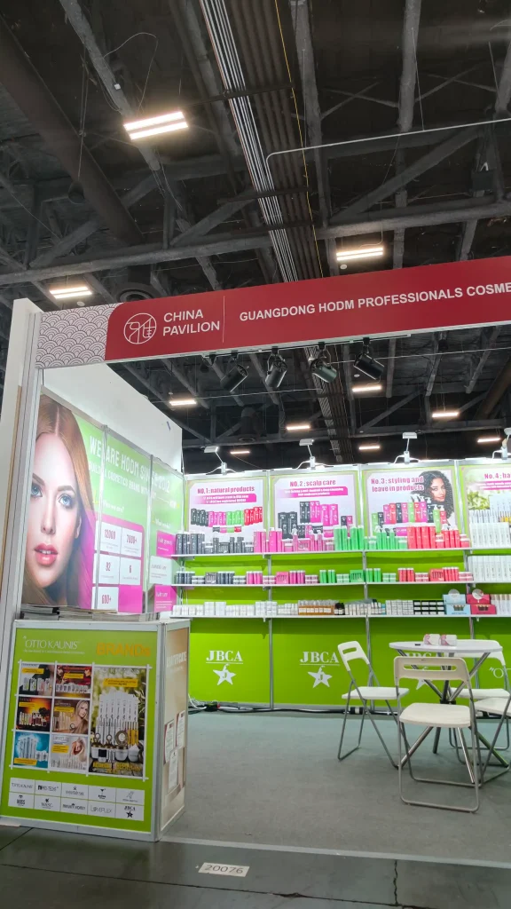 Booth at a trade show labeled 'China Pavilion' with cosmetics products on bright green and pink shelves and display boards behind. A few white chairs are nearby under a high ceiling with exposed beams.