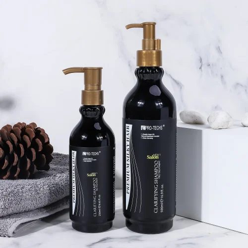 Two black bottles of "CLARIFYING SHAMPOO" in 250ml and 500ml sizes with gold pump dispensers, set on a marble surface alongside a pinecone, a grey towel, and white decorative stones.