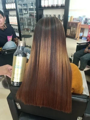 Long, straight hair with caramel and dark brown balayage resting on a salon chair, with a gold-labeled hair product bottle in the foreground.