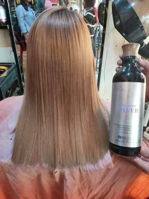 Back view of person with shiny straight blonde hair in a salon, holding a silver protein bottle used for hair treatment behind them.