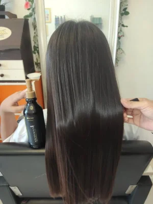 Rear view of a person with long, glossy straight dark hair being touched by a hand, next to a black keratin treatmnet bottle in a salon