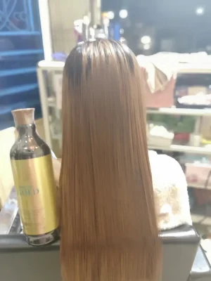 Rear view of a woman with long, shiny straight brown hair next to a glod keratin treatment bottle in a salon setting